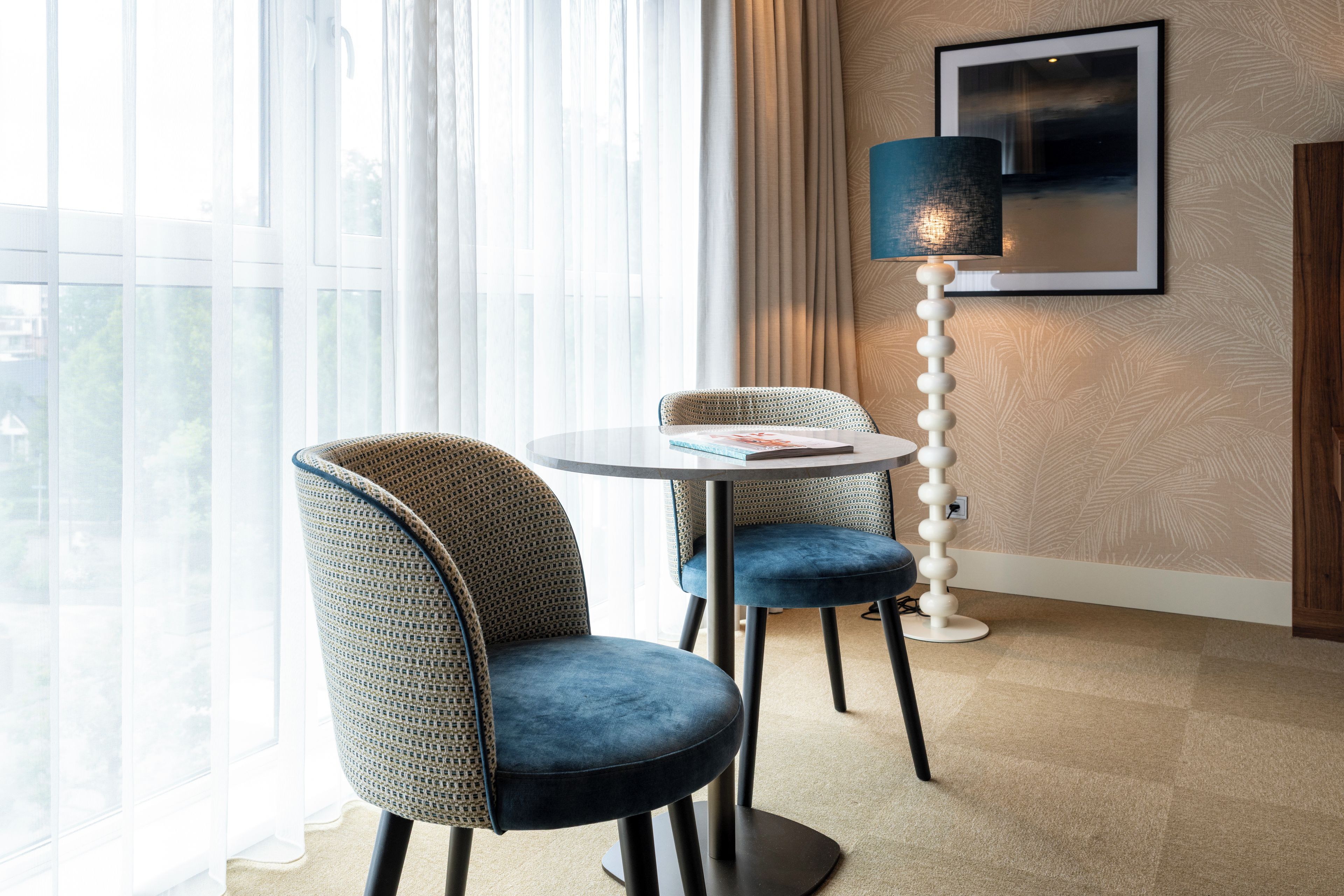A modern hotel room with a round table, two upholstered chairs, a floor lamp, and a large window with sheer curtains.