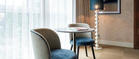 A modern hotel room with a round table, two upholstered chairs, a floor lamp, and a large window with sheer curtains.