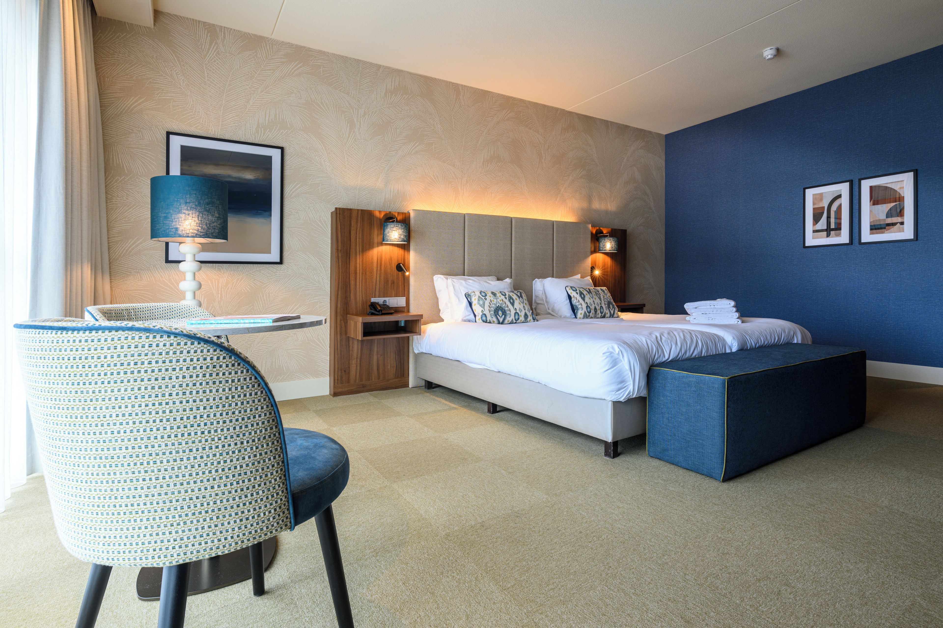 A hotel room with a bed, bedside table, lamp, chair, and artwork on the wall.