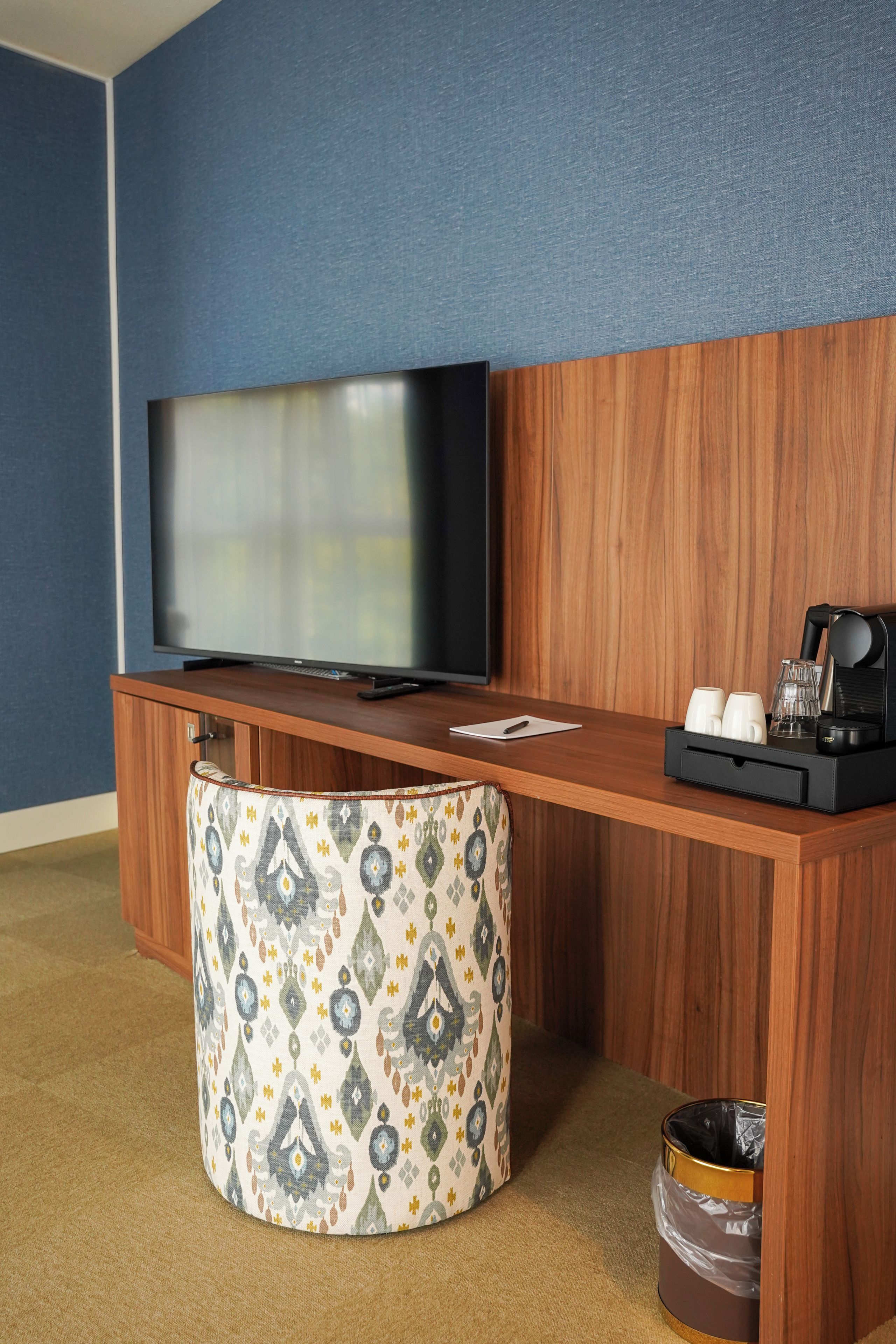A hotel room with a wooden desk, a television, a patterned ottoman, and a coffee maker.