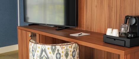 A hotel room with a wooden desk, a television, a patterned ottoman, and a coffee maker.