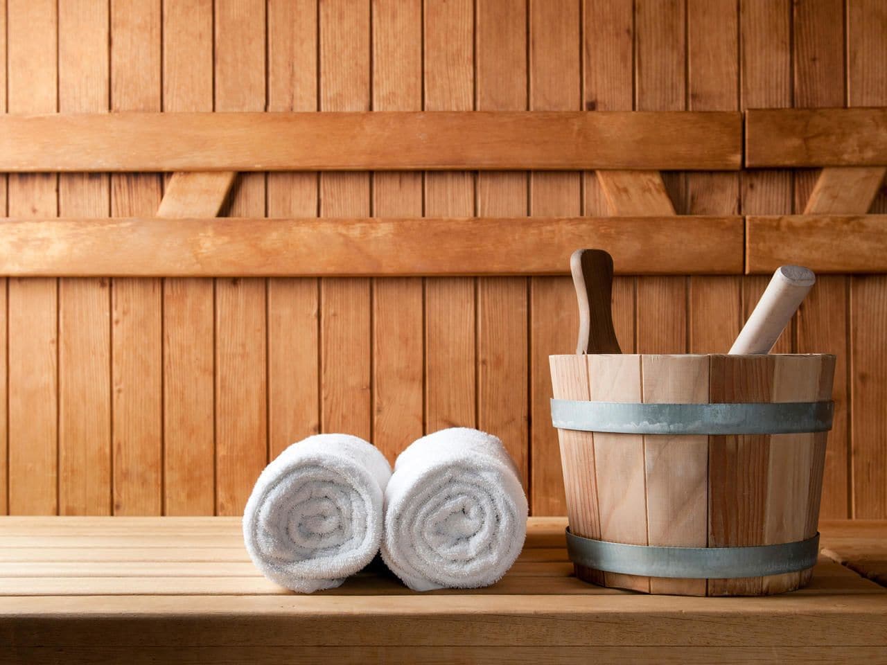 Een houten sauna bank met twee opgerolde handdoeken en een houten emmer met een houten hamer.