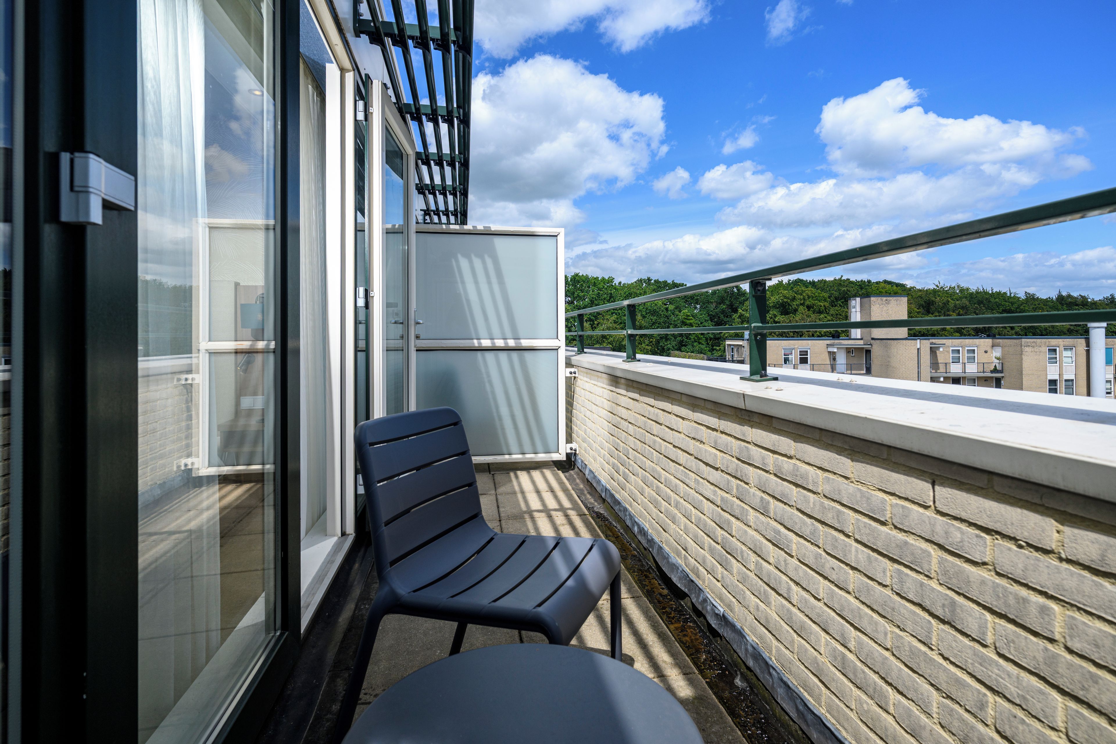 A balcony with a chair and a table, a glass door, and a view of buildings and trees.