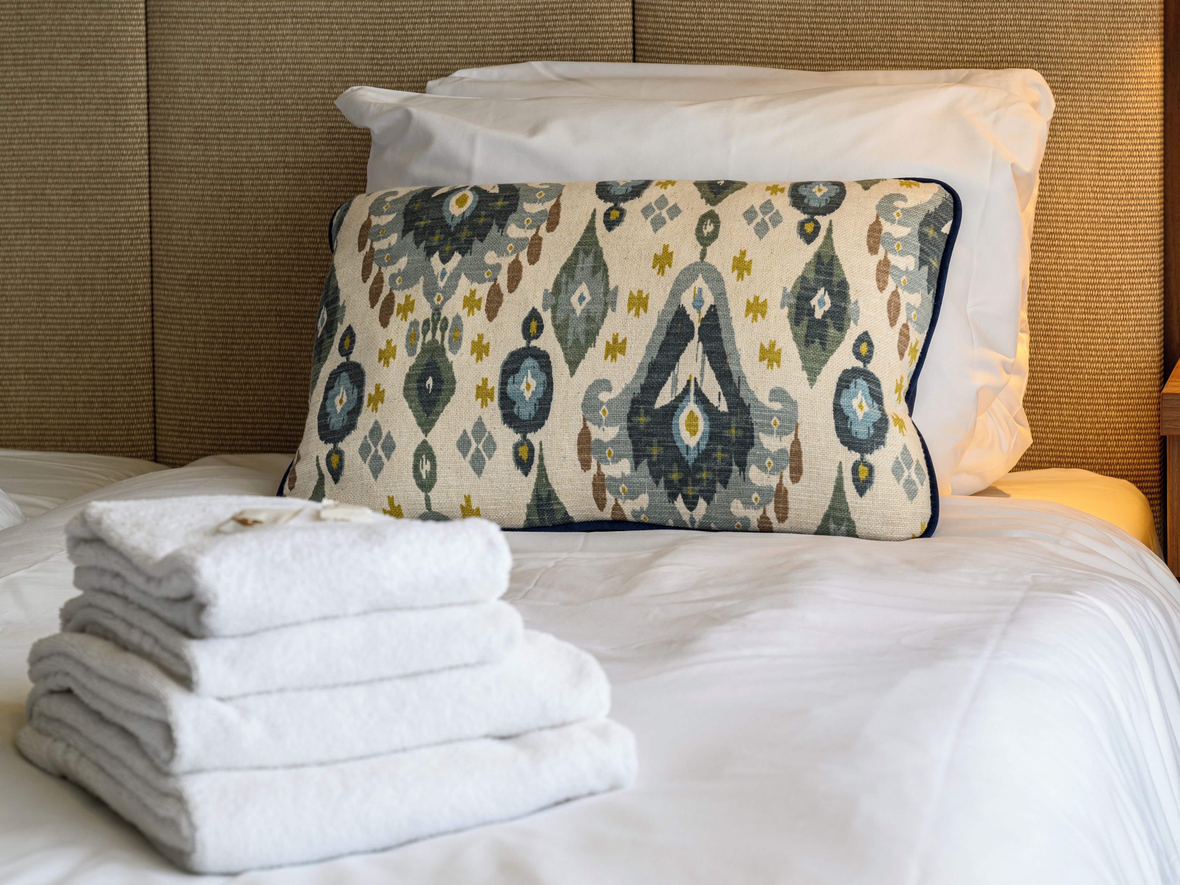 Cama bem arrumada com lençóis brancos, almofada estampada e uma pilha de toalhas dobradas.