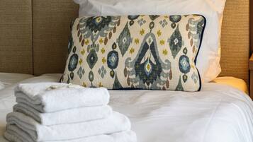 Cama bem arrumada com lençóis brancos, almofada estampada e uma pilha de toalhas dobradas.