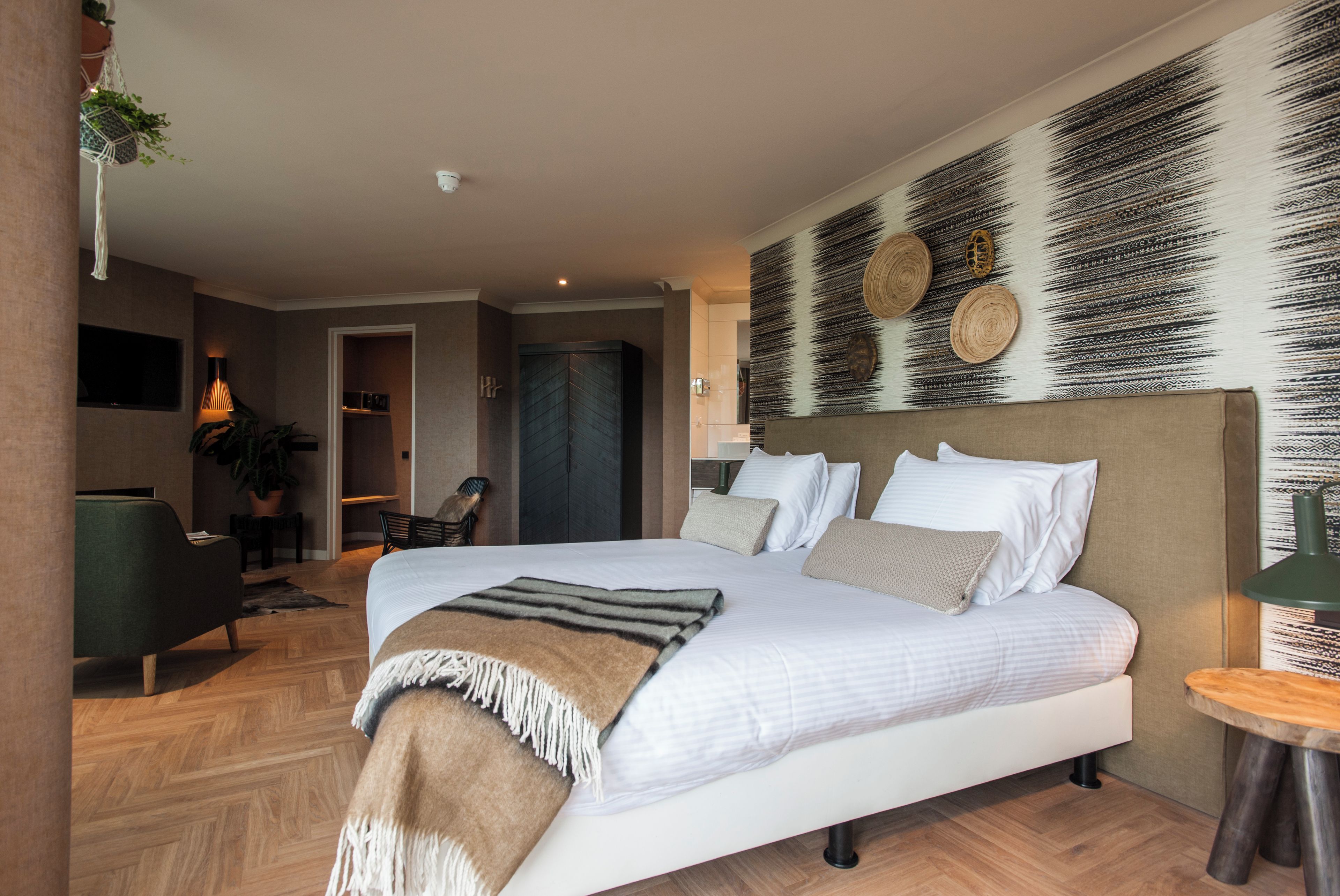A modern hotel room with a large bed, a flat-screen TV, a seating area with a chair and table, and a decorative wall.