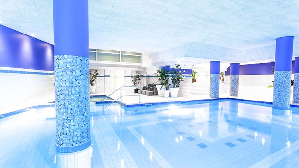 A large indoor swimming pool with blue-tiled walls and columns, surrounded by seating areas and potted plants.