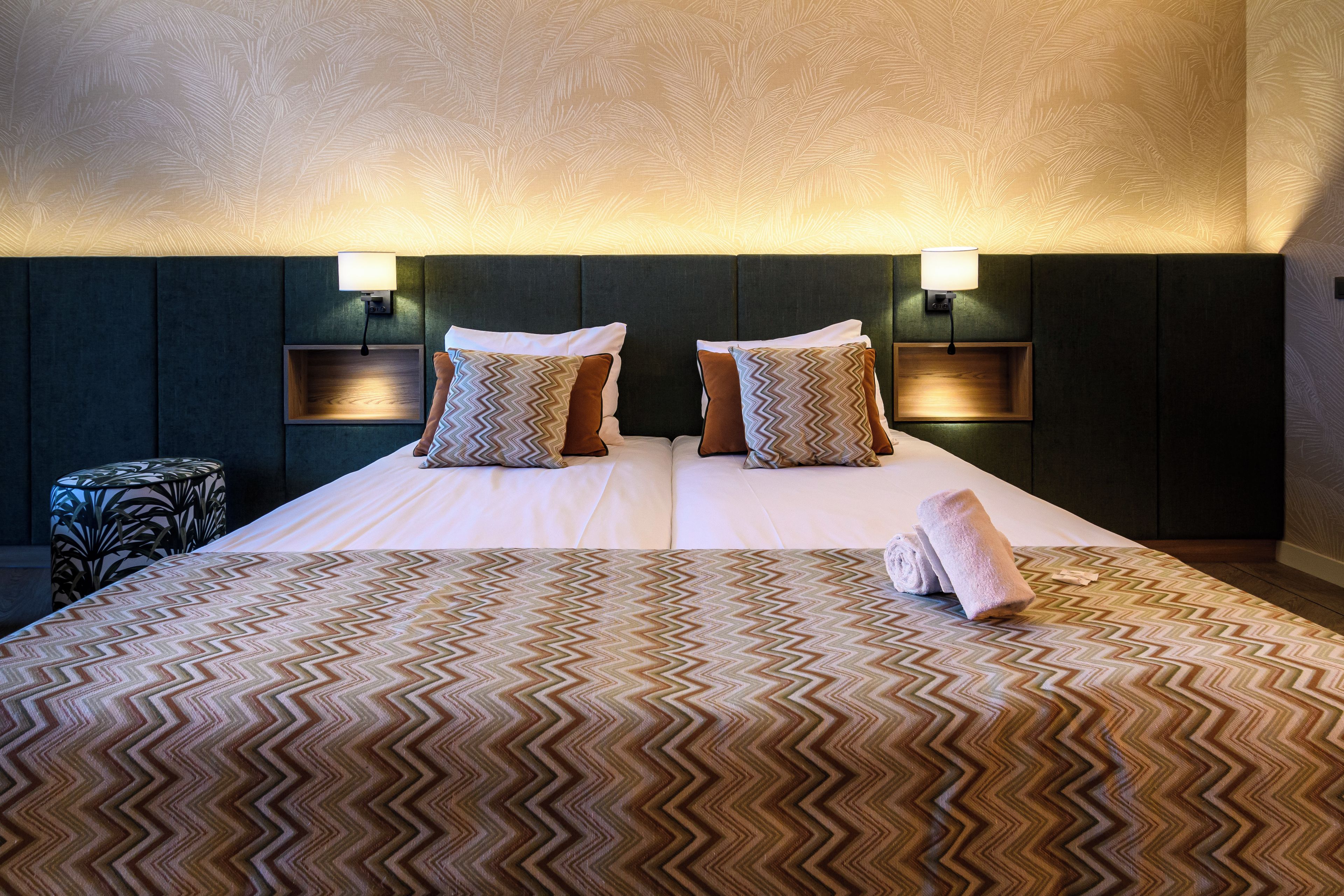 Habitación de hotel con una cama grande, dos almohadas, cabecero con luces empotradas, una mesita redonda y una toalla enrollada sobre la cama.