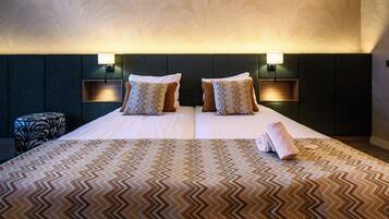 Habitación de hotel con una cama grande, dos almohadas, cabecero con luces empotradas, una mesita redonda y una toalla enrollada sobre la cama.