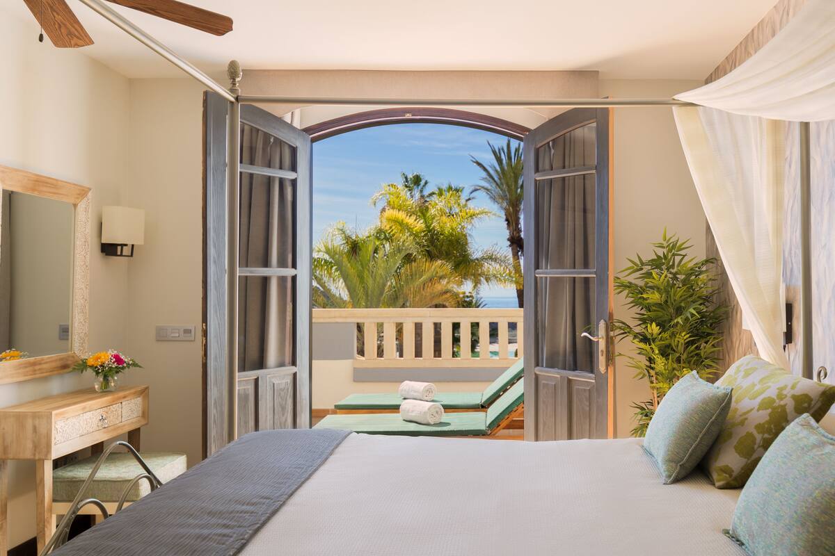 A hotel room with a bed, a vanity with a mirror, a small table with a towel, and a view of the outdoors.