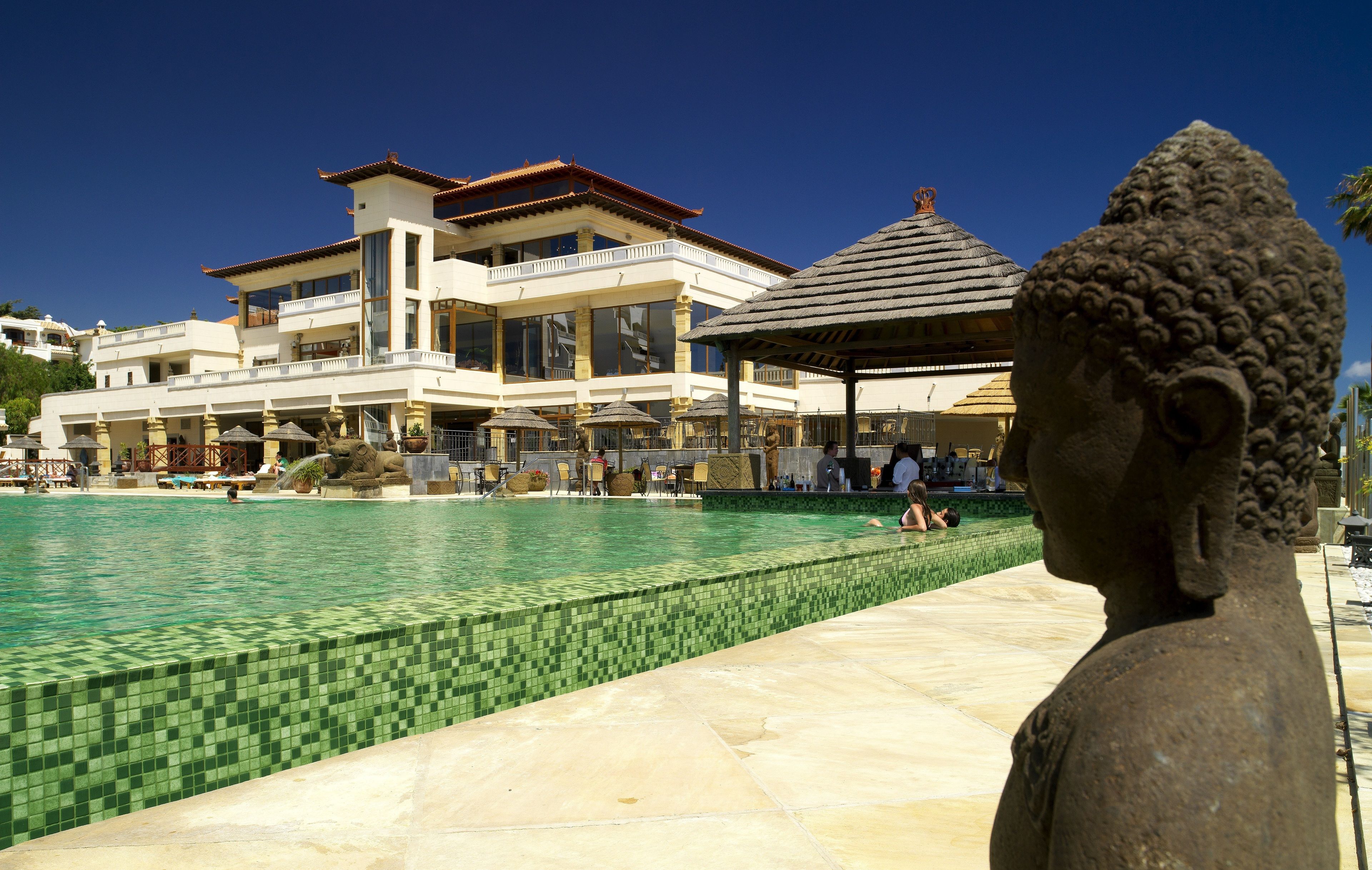 Zona de piscina con una estatua de Buda, un edificio grande con balcones y un borde de piscina con azulejos.