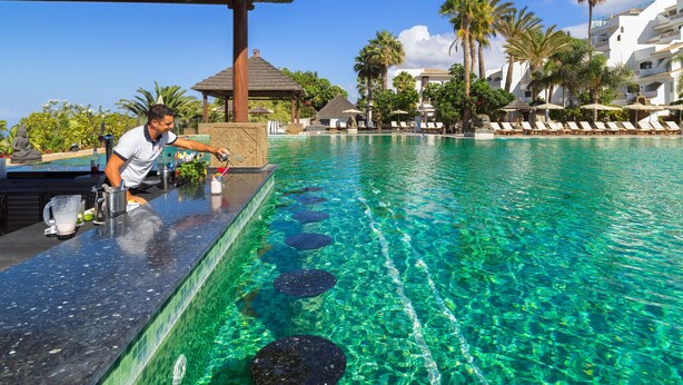 A poolside bar with a bartender, clear blue water, and lounge chairs.