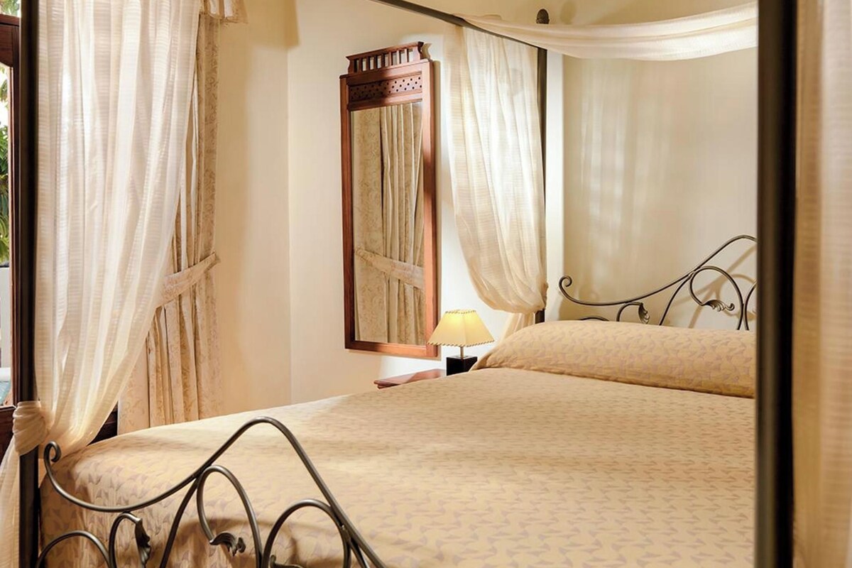 Una cama con dosel, una mesita de noche con lámpara y cortinas.