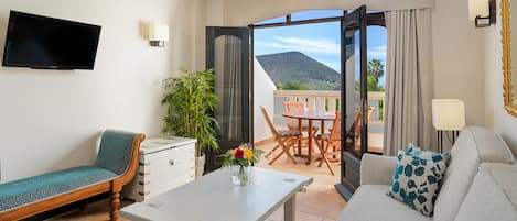 A modern living room with a sofa, a dining table, and a TV. There is a balcony with a table and chairs, and a large window offering a view of the mountains.