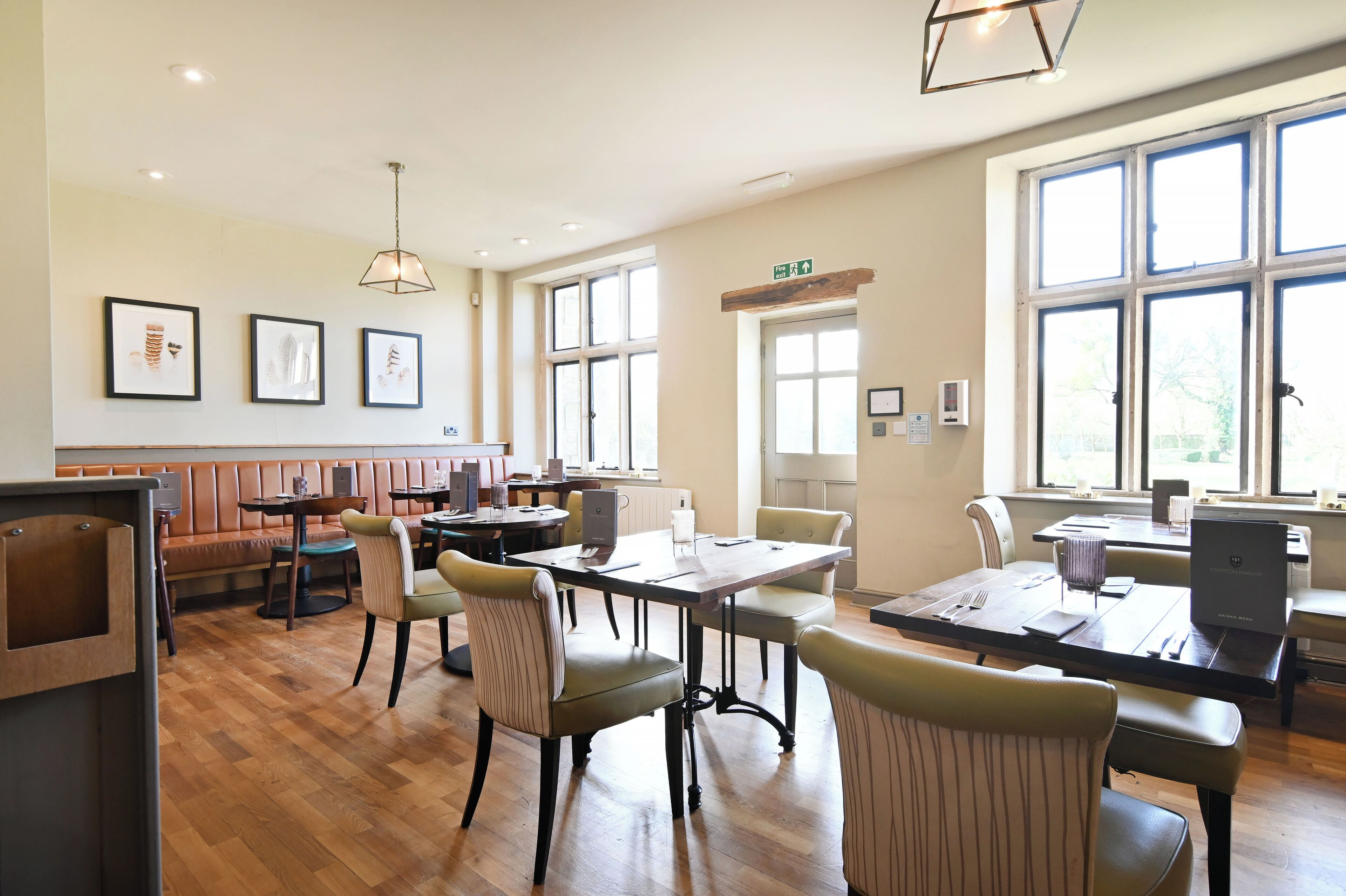 A spacious dining area with wooden flooring, multiple tables and chairs, framed artwork on the walls, and large windows providing natural light.