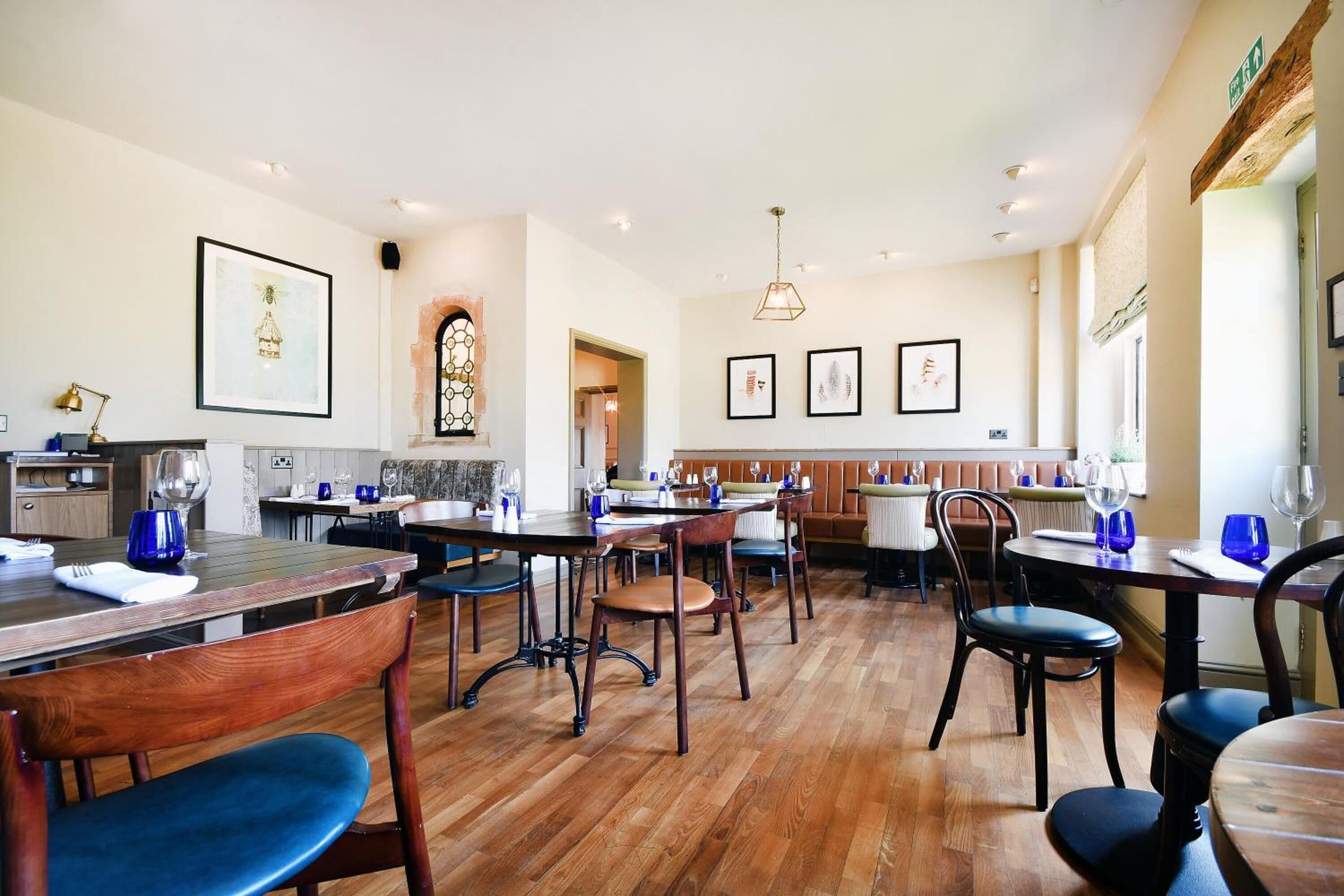 A dining area with wooden tables and chairs, a kitchen in the background, and framed artwork on the walls.