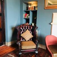 A dark brown leather armchair with a plaid cushion in a room with blue walls, wooden flooring, and a bookshelf in the background.