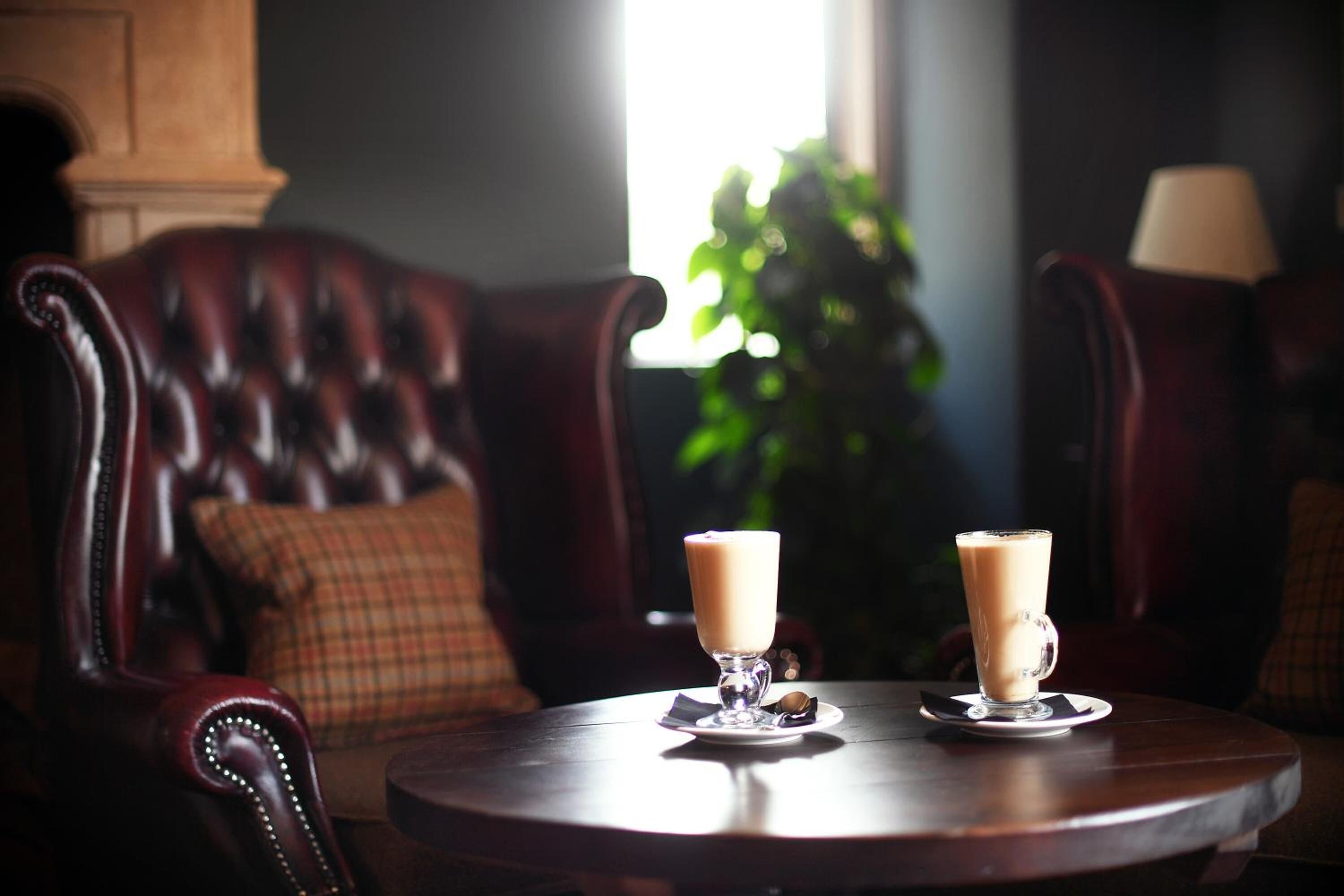 Una mesa redonda de madera con dos vasos de una bebida clara, probablemente café o té, en un vaso con platillo. La mesa está frente a un sofá de cuero oscuro con un cojín a cuadros.