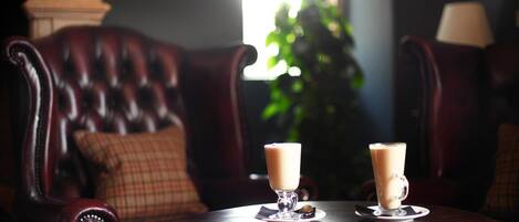 Una mesa redonda de madera con dos vasos de una bebida clara, probablemente café o té, en un vaso con platillo. La mesa está frente a un sofá de cuero oscuro con un cojín a cuadros.