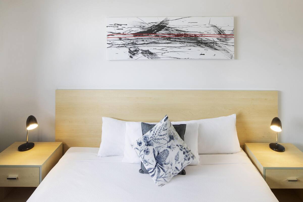A hotel room with a bed, two bedside tables with lamps, a wooden headboard, and a wall-mounted abstract painting.