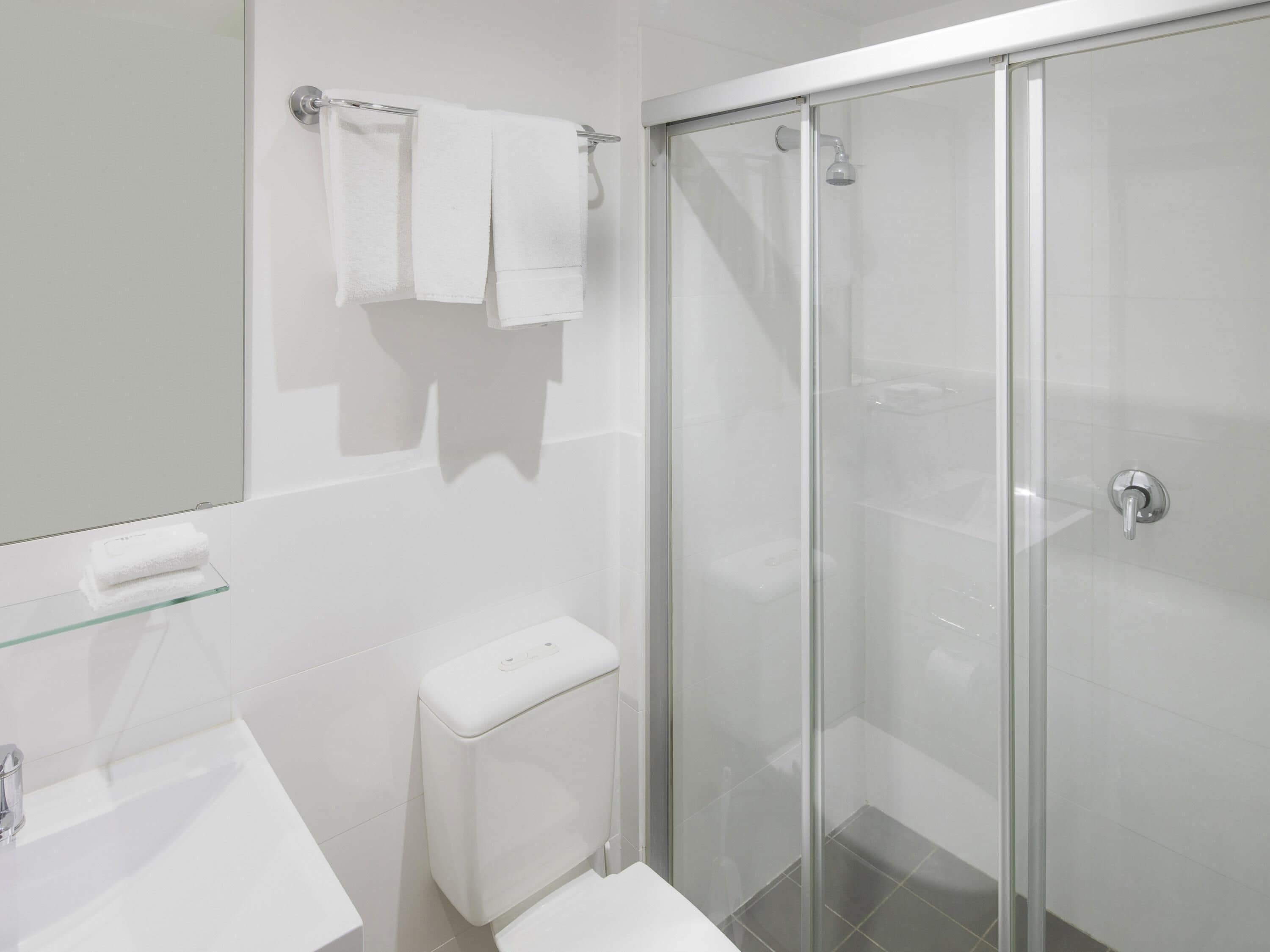 Une salle de bain moderne avec une douche à parois de verre, des toilettes blanches et une étagère en verre sur laquelle est posée une serviette.