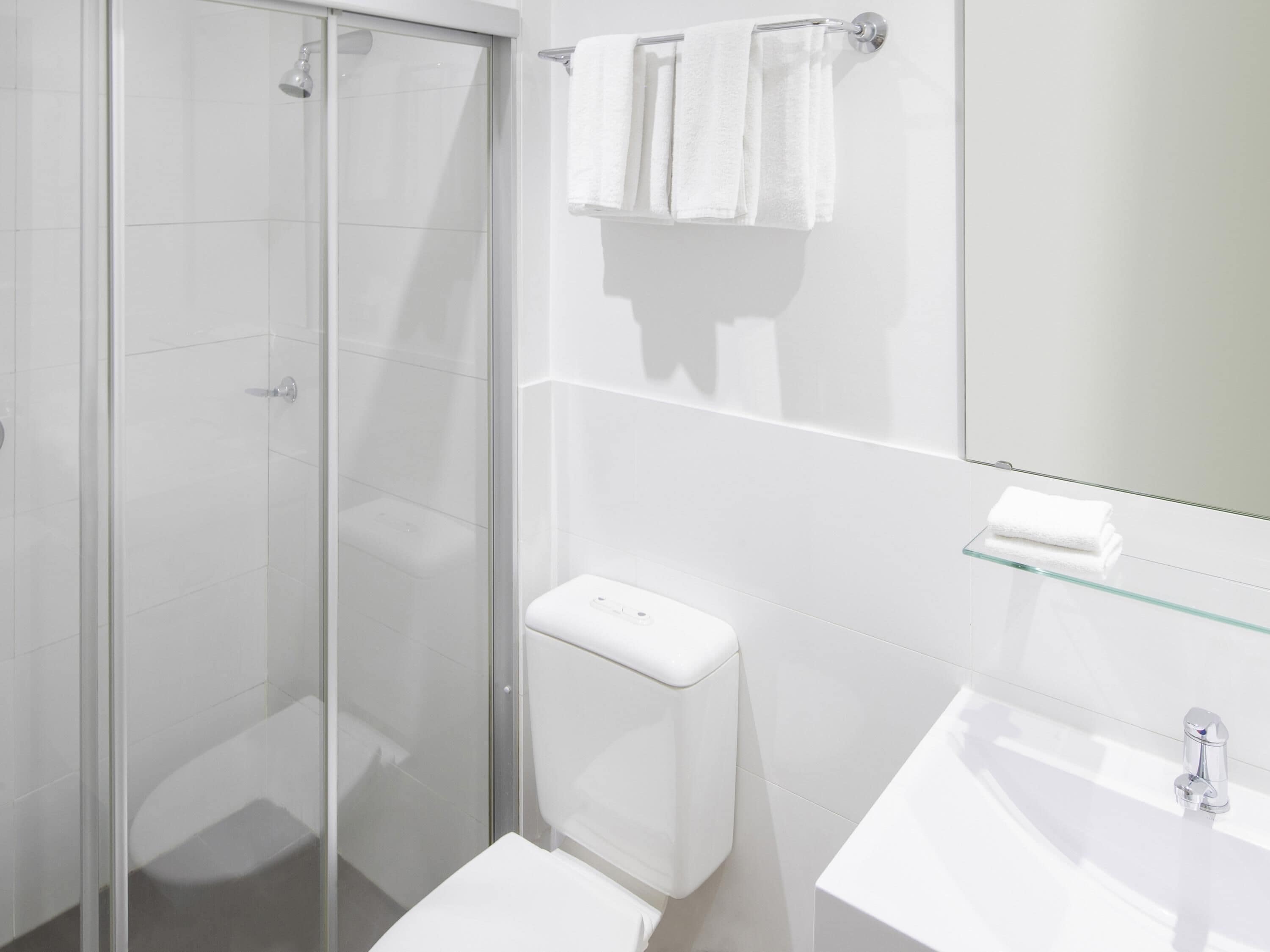 Un baño moderno con cabina de ducha de vidrio, un inodoro blanco, un estante de vidrio con una toalla y un lavabo con grifo cromado.