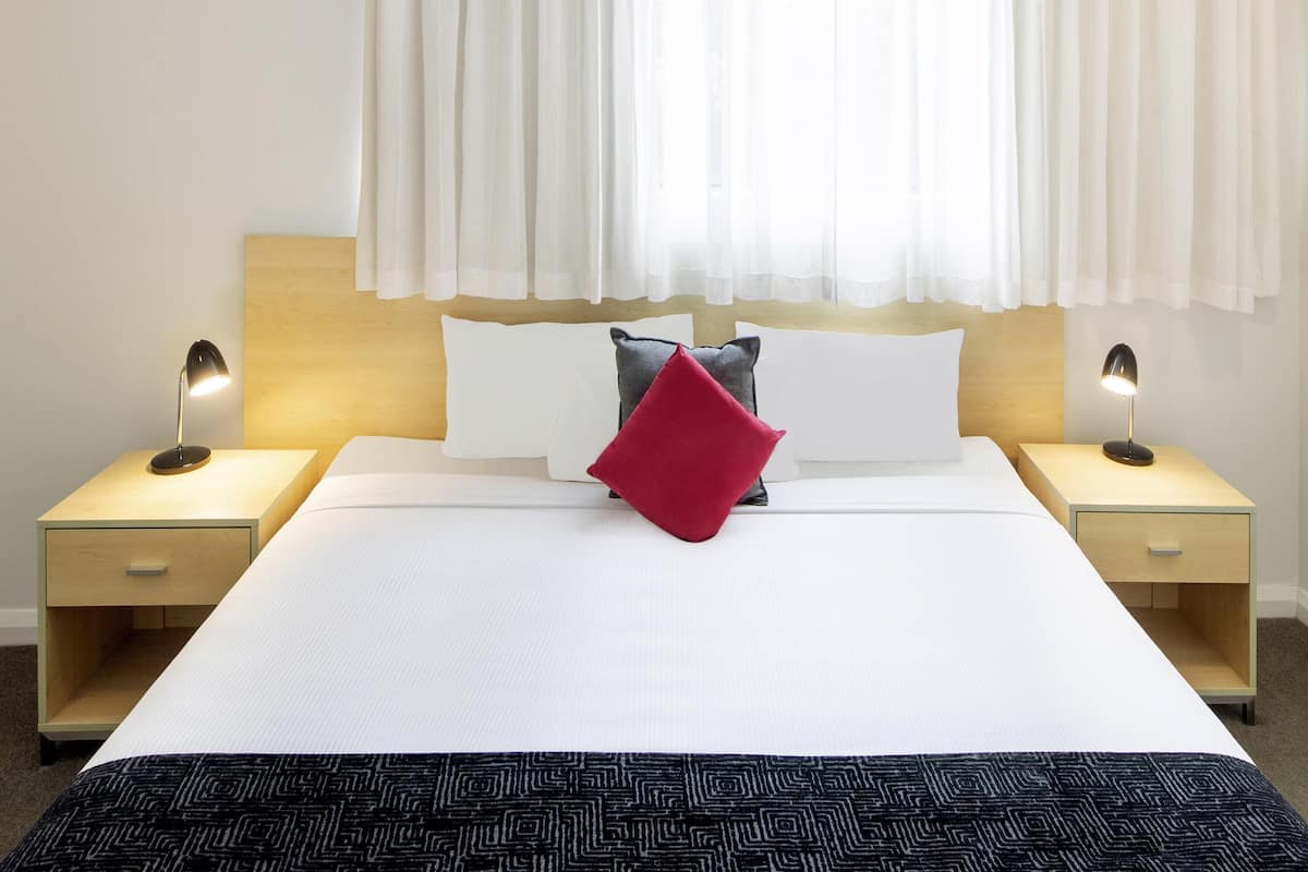 A hotel room with a large bed, two bedside tables, a nightstand, a wooden headboard, and a window with sheer curtains.