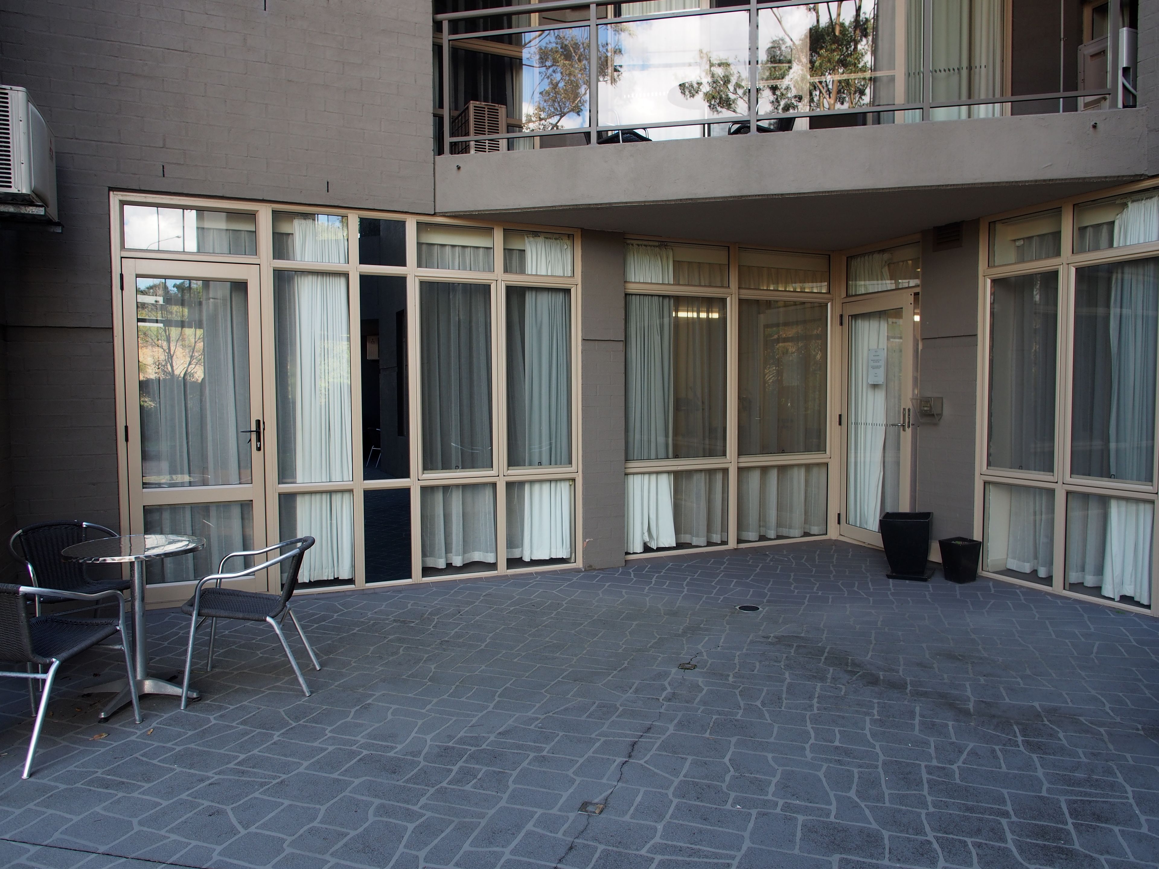 Pátio com mesa e cadeiras, janelas amplas com cortinas e uma lixeira.