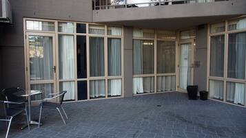 A courtyard with a patio table and chairs, large windows with curtains, and a trash bin.