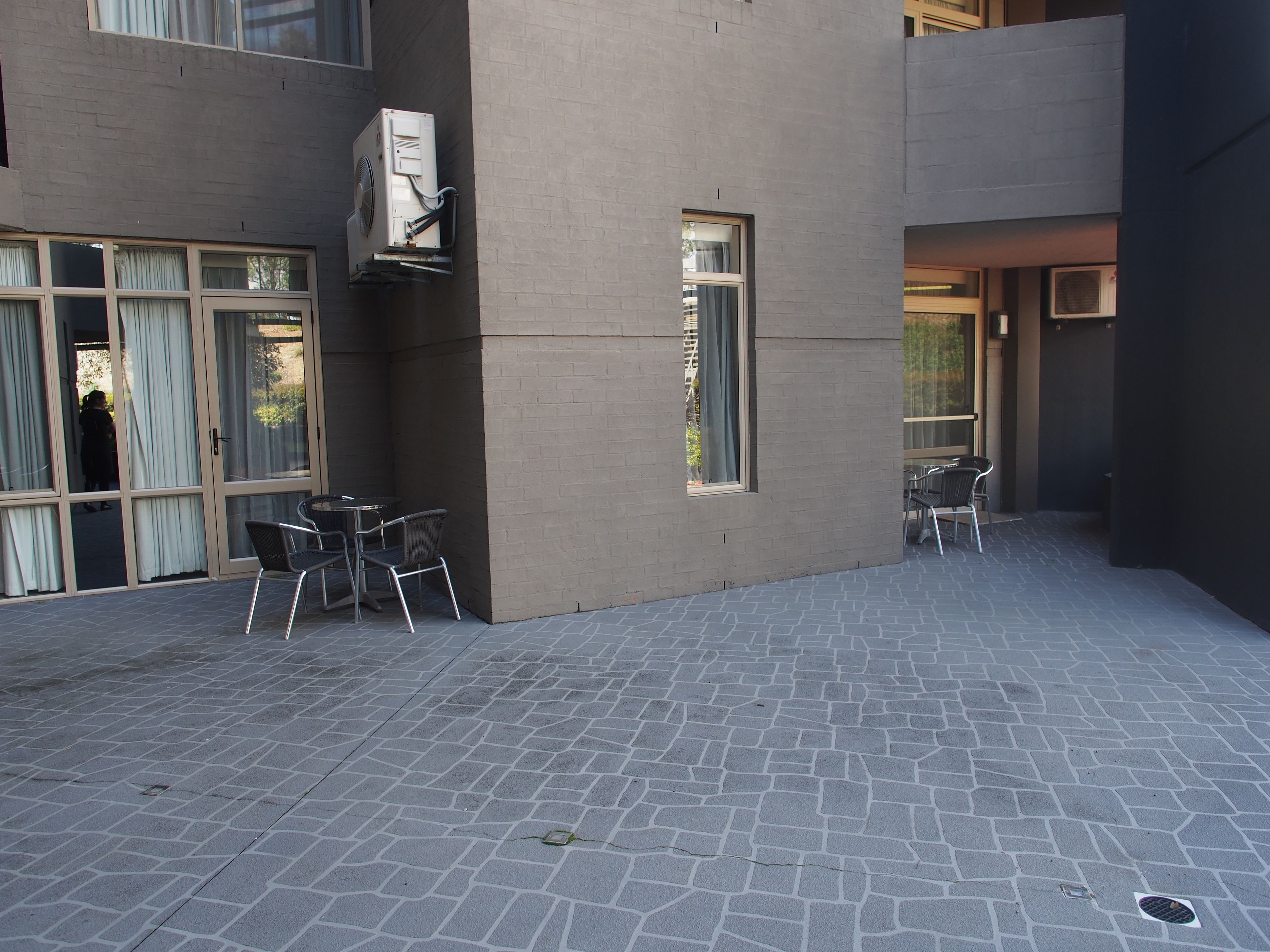 Un espace extérieur aménagé en terrasse, avec un sol en pierre, deux tables accompagnées de chaises et une grande fenêtre.