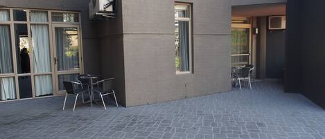 Un espace extérieur aménagé en terrasse, avec un sol en pierre, deux tables accompagnées de chaises et une grande fenêtre.