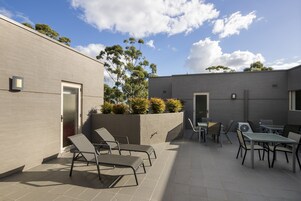 Un patio moderno al aire libre con sillones y mesas, un edificio gris y un cielo despejado.