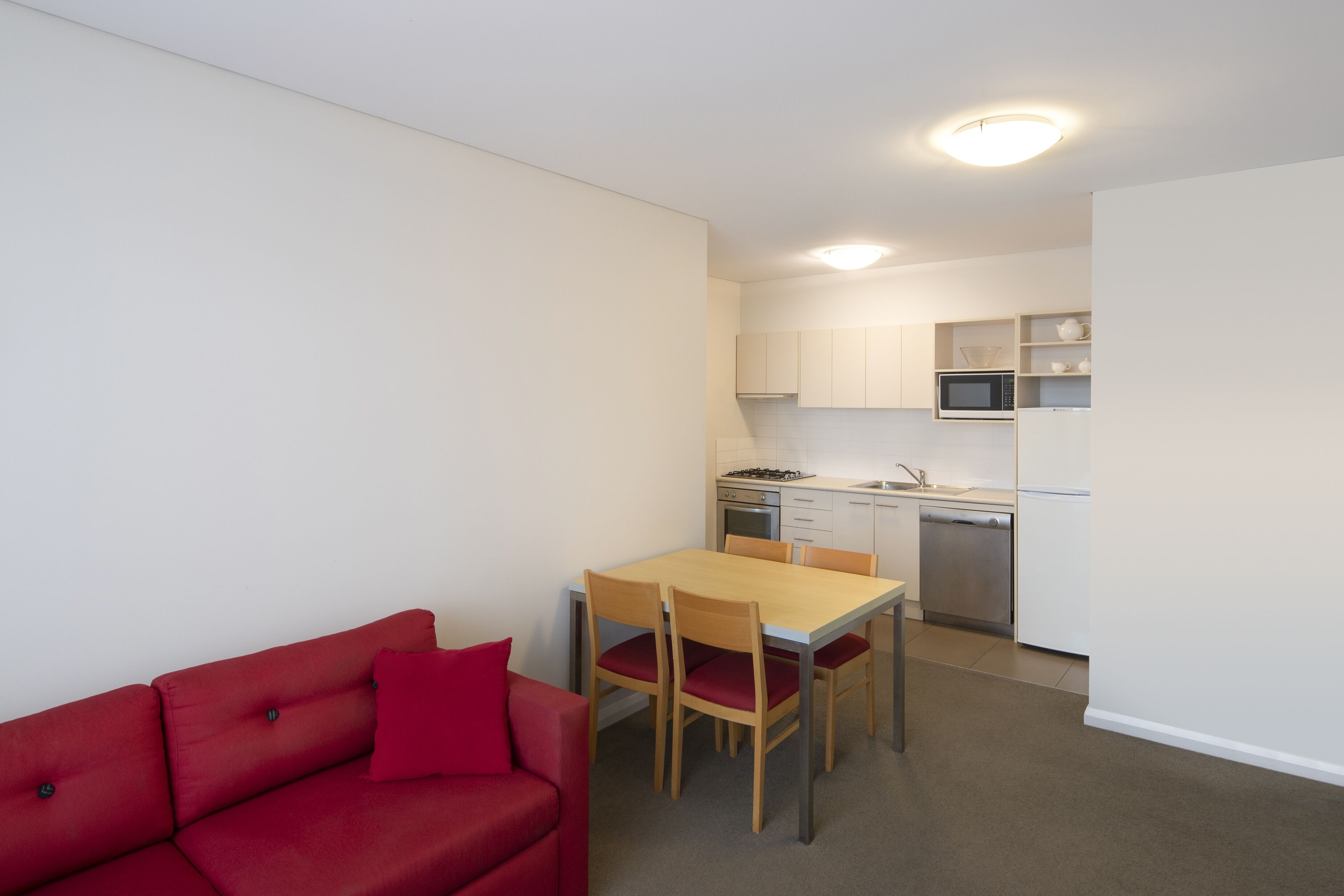 A compact living space with a red sofa, a wooden dining table with four chairs, a kitchen area with white cabinets, a microwave, a stove, and a dishwasher.