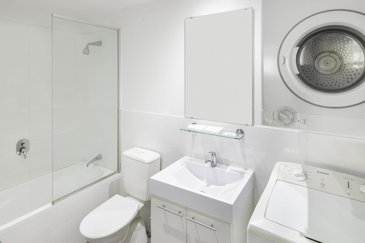 A modern bathroom with a glass shower enclosure, a toilet, a sink with a mirror, and a front-loading washing machine.