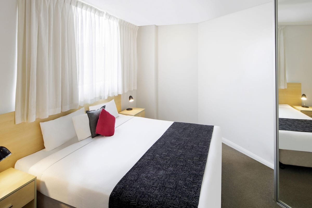 A hotel room with a bed, a nightstand with a lamp, a mirror, and a window with curtains.