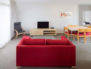 Ein modernes Wohnzimmer mit einem roten Sofa, einem Fernseher auf einem Holzkasten, einem Esstisch mit Stühlen und einem Fenster mit weißen Vorhängen.