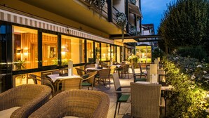 Un área de comedor al aire libre con mobiliario de mimbre y mesas, rodeada por un edificio con ventanas grandes y macetas colgantes de flores.