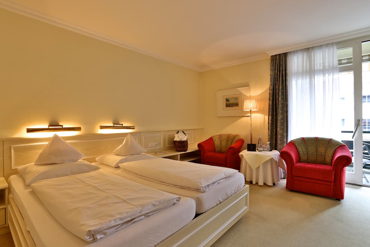 A hotel room with two beds, a red armchair, a small table, and a window with curtains.