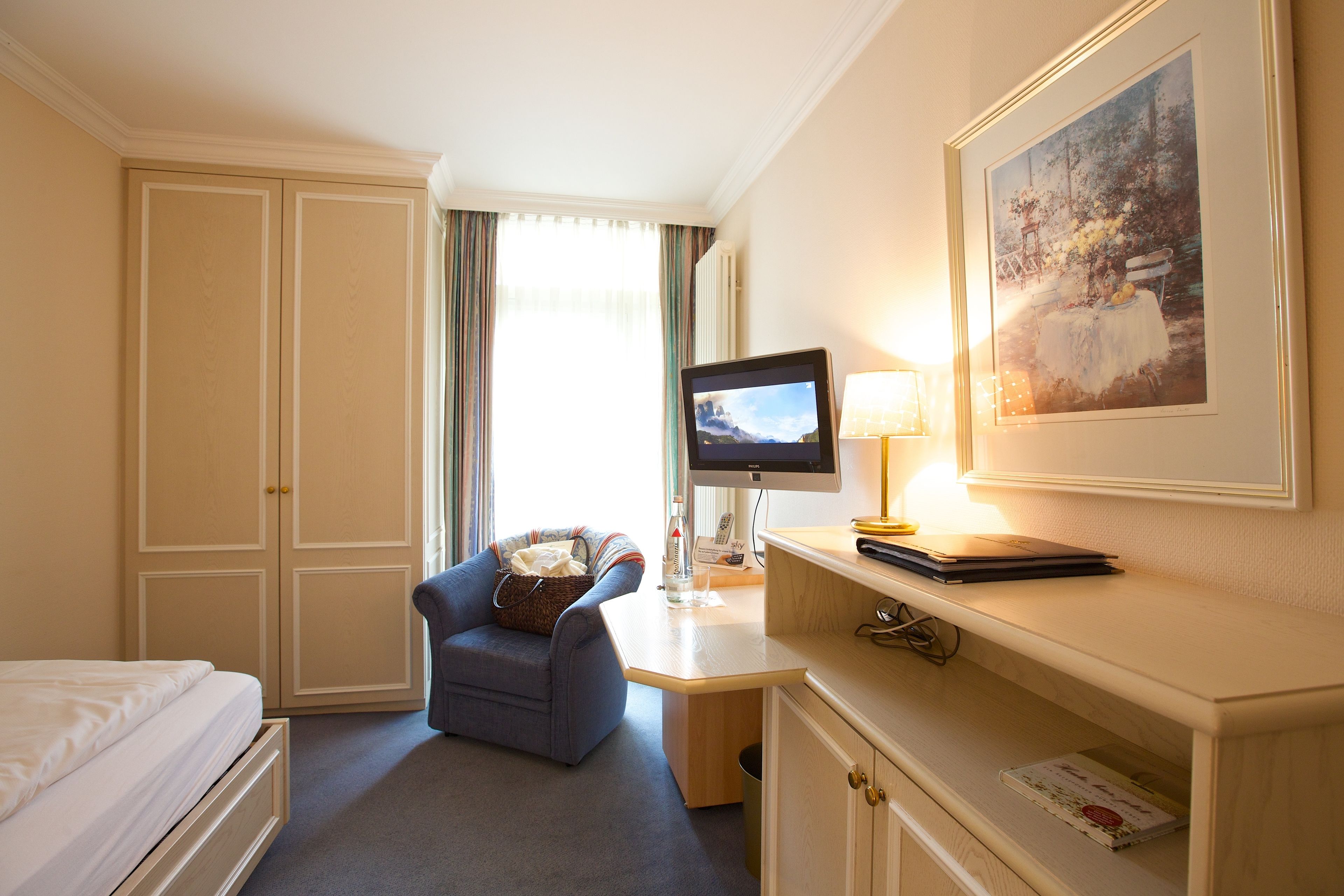 A hotel room with a bed, a desk with a computer, a chair, a lamp, and a framed picture on the wall.
