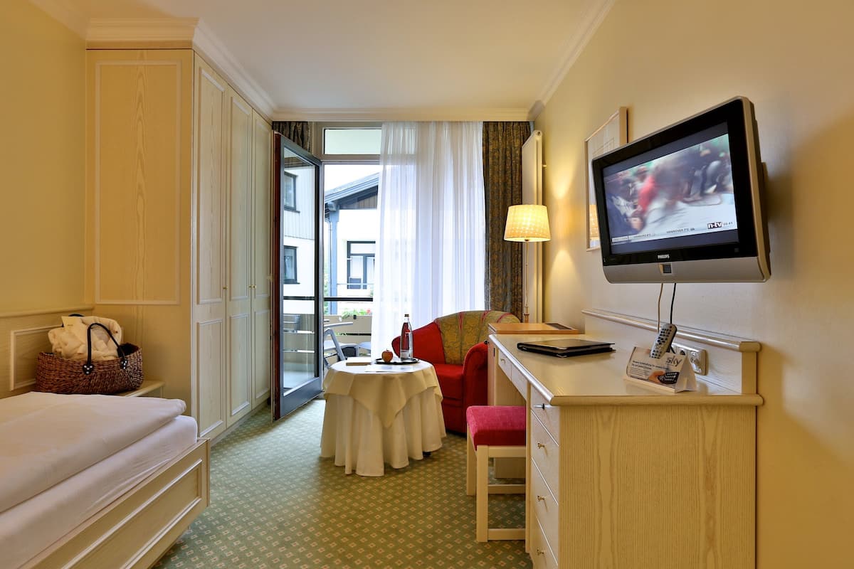 A hotel room with a bed, a desk with a television, a small table, a chair, and a window with curtains.