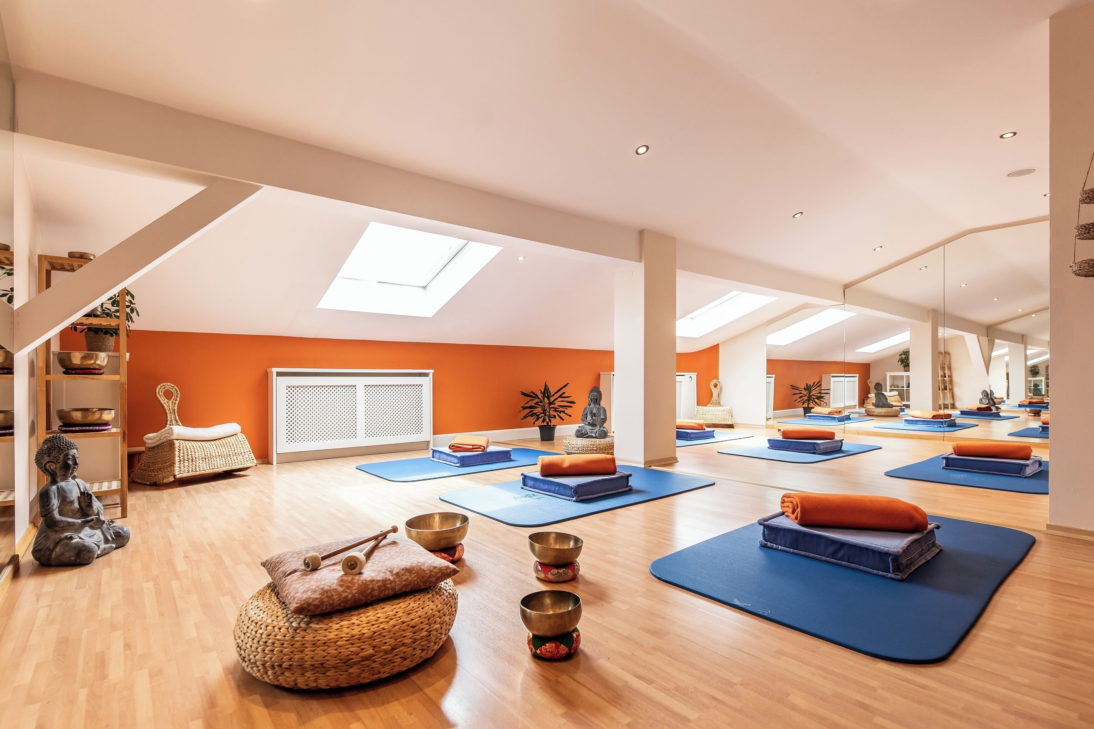 Ruime kamer met houten vloeren, oranje wanden en een dakraam. Er staan yogamatten, meditatiekussens en een beeld.