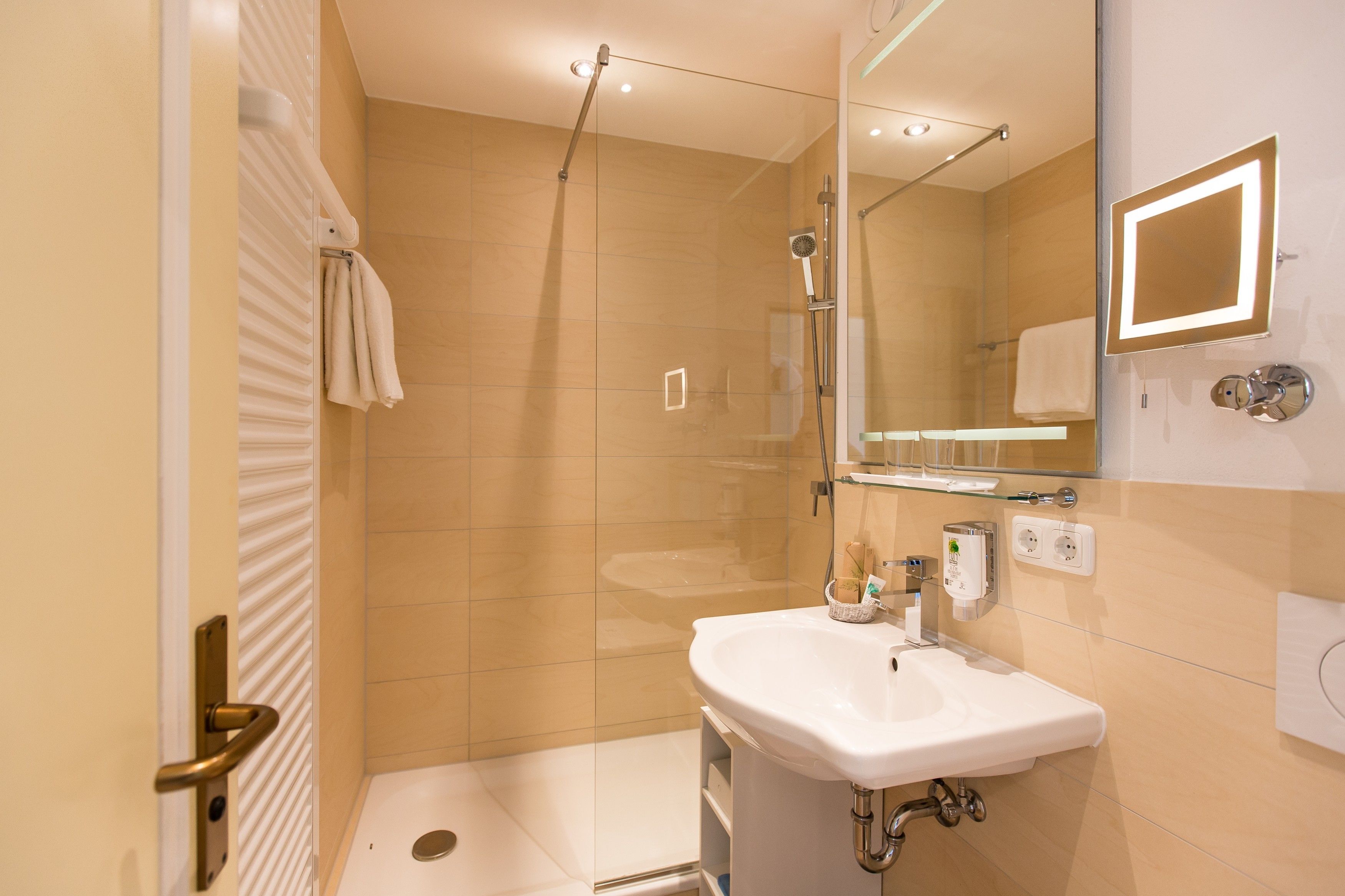 Un bany modern amb un compartiment de dutxa de vidre, un lavabo blanc i un mirall amb il·luminació incrustada.