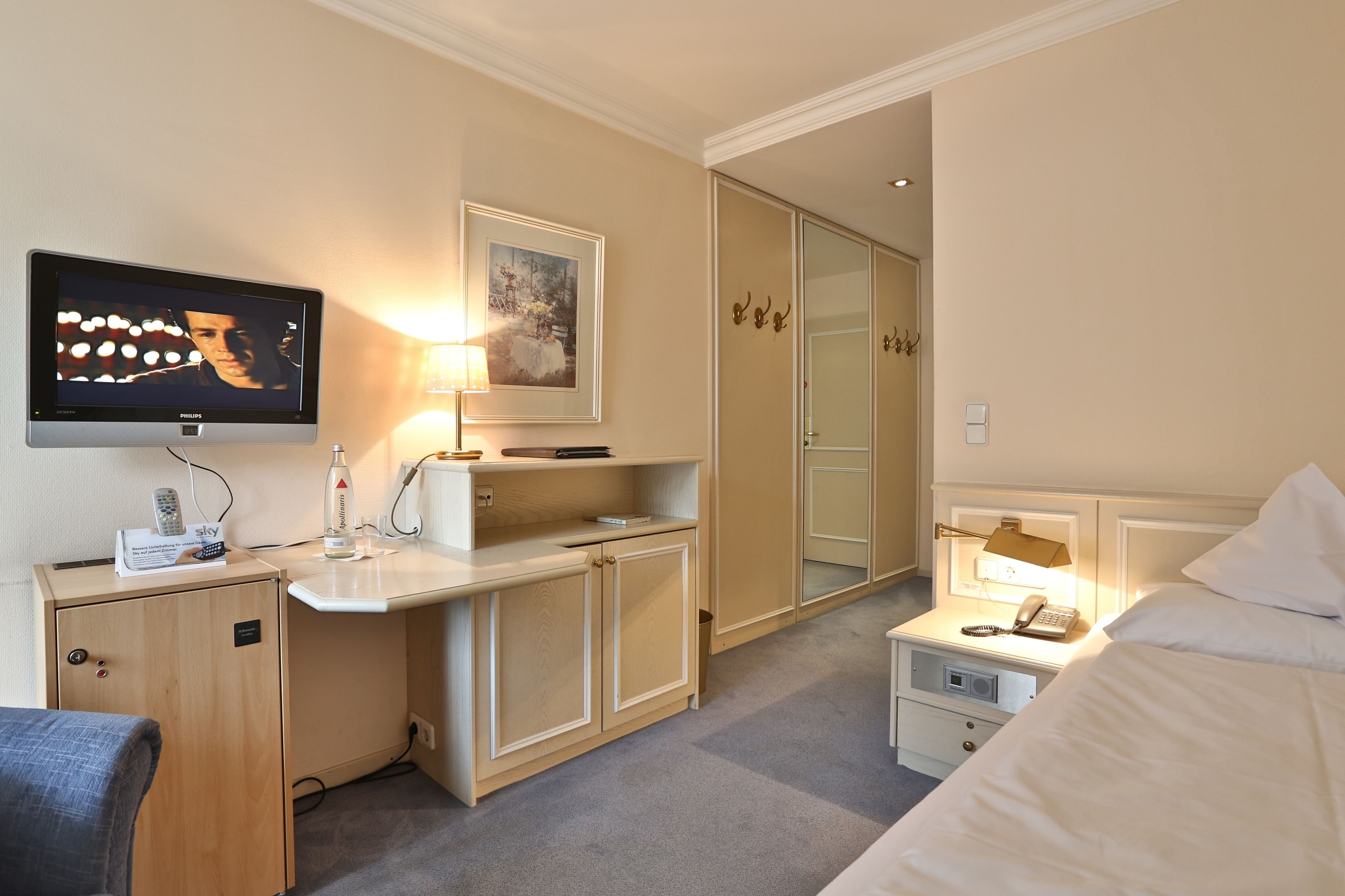 A hotel room with a television, bedside lamp, a desk with a lamp, a bed, a wardrobe, and a mirror.