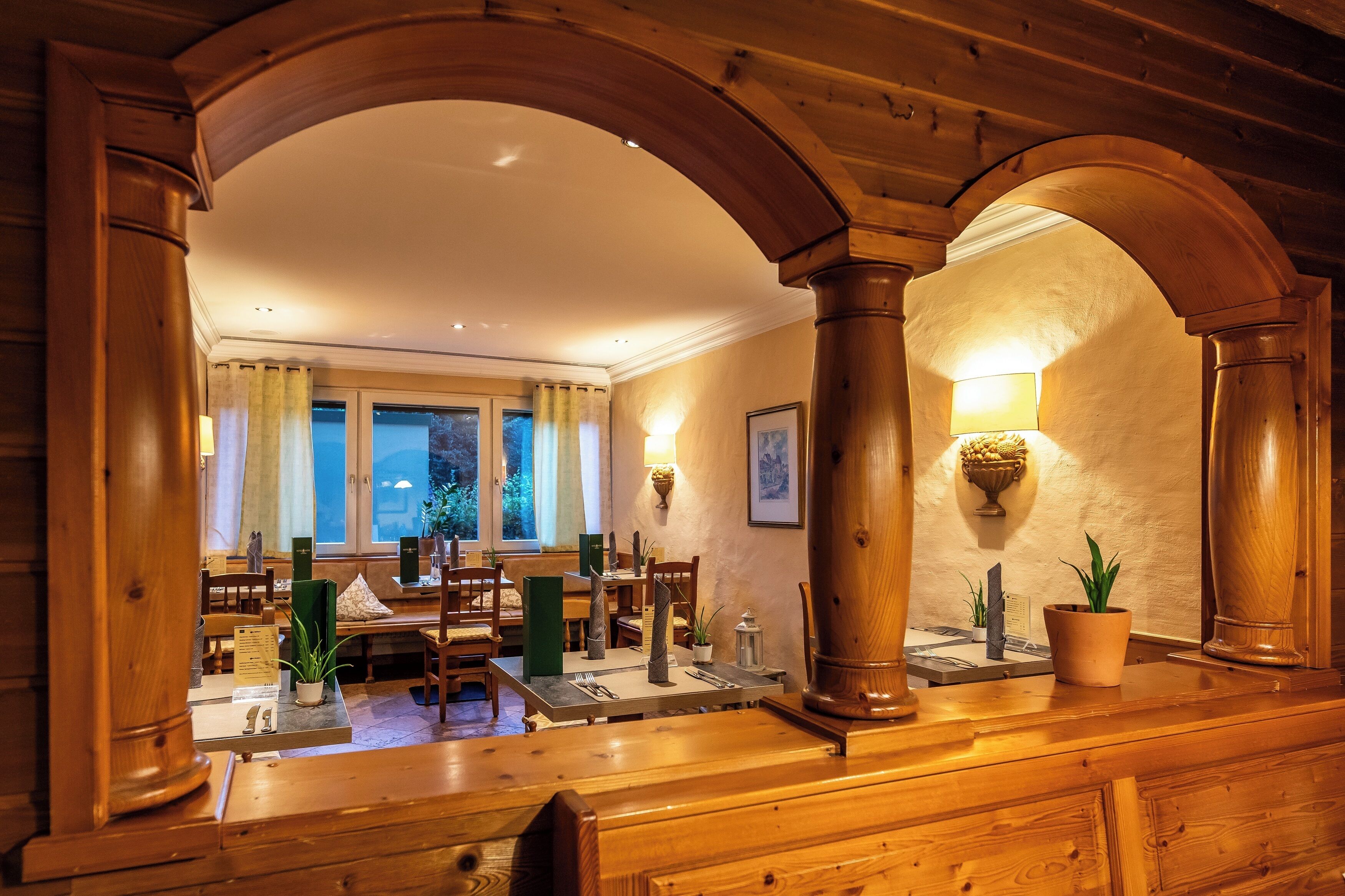 A warmly lit dining area with wooden furniture, a large window with curtains, and a wall-mounted lamp.