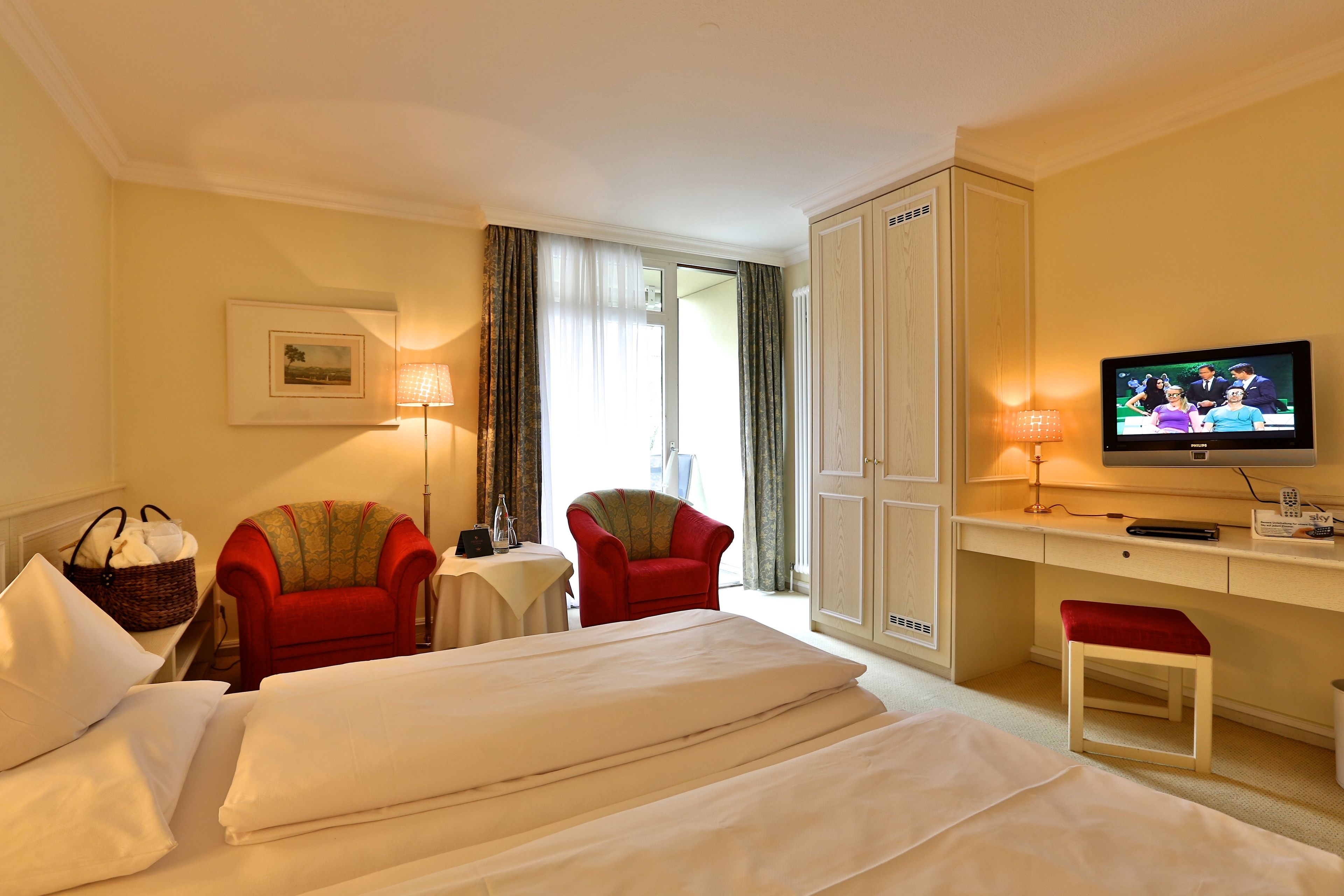 Ein Hotelzimmer mit einem Bett, zwei roten Sesseln, einem Schreibtisch mit Fernseher und einem großen Kleiderschrank.