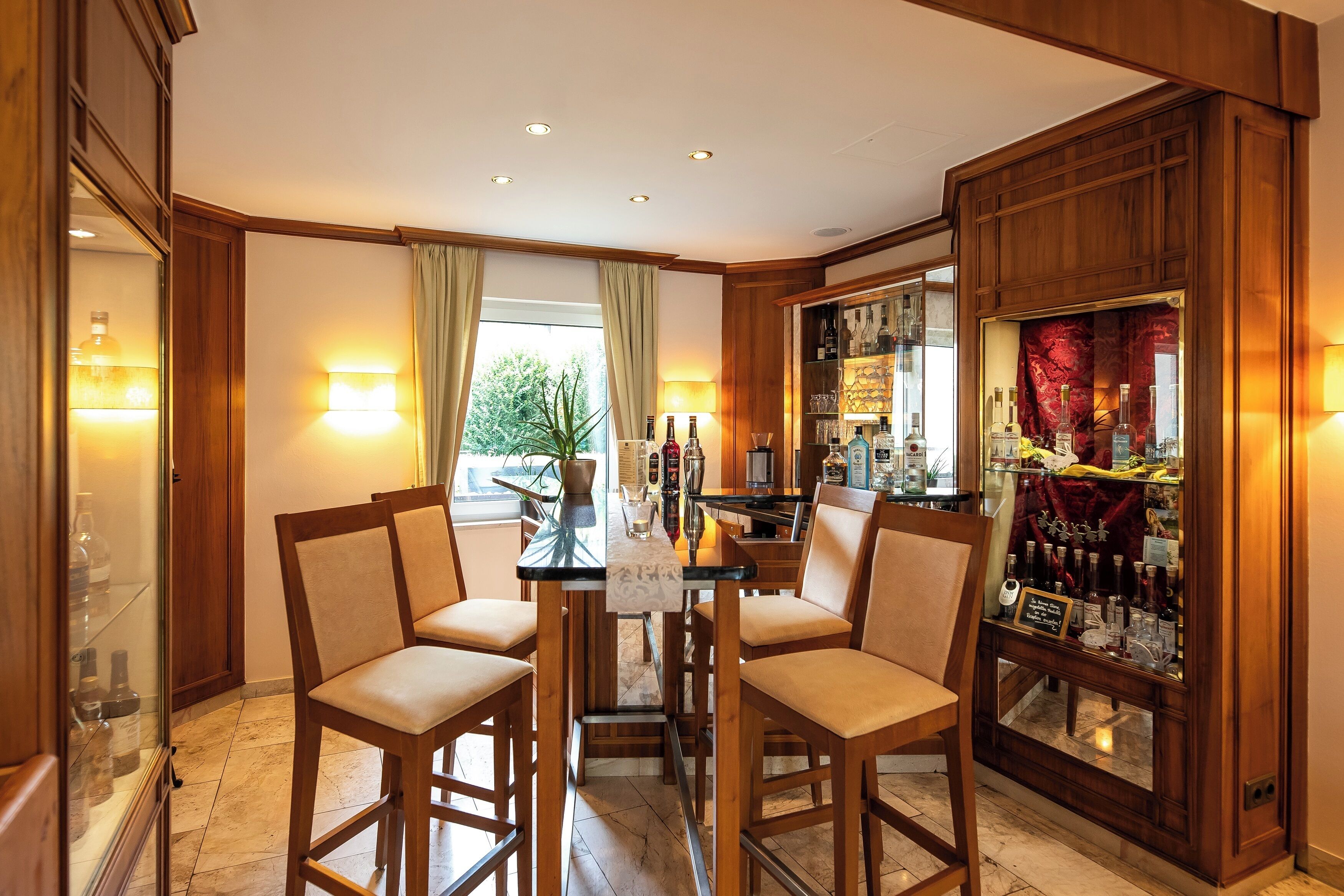 A dining area with a table set for four, surrounded by wooden chairs. A bar with a glass display case is visible to the right.