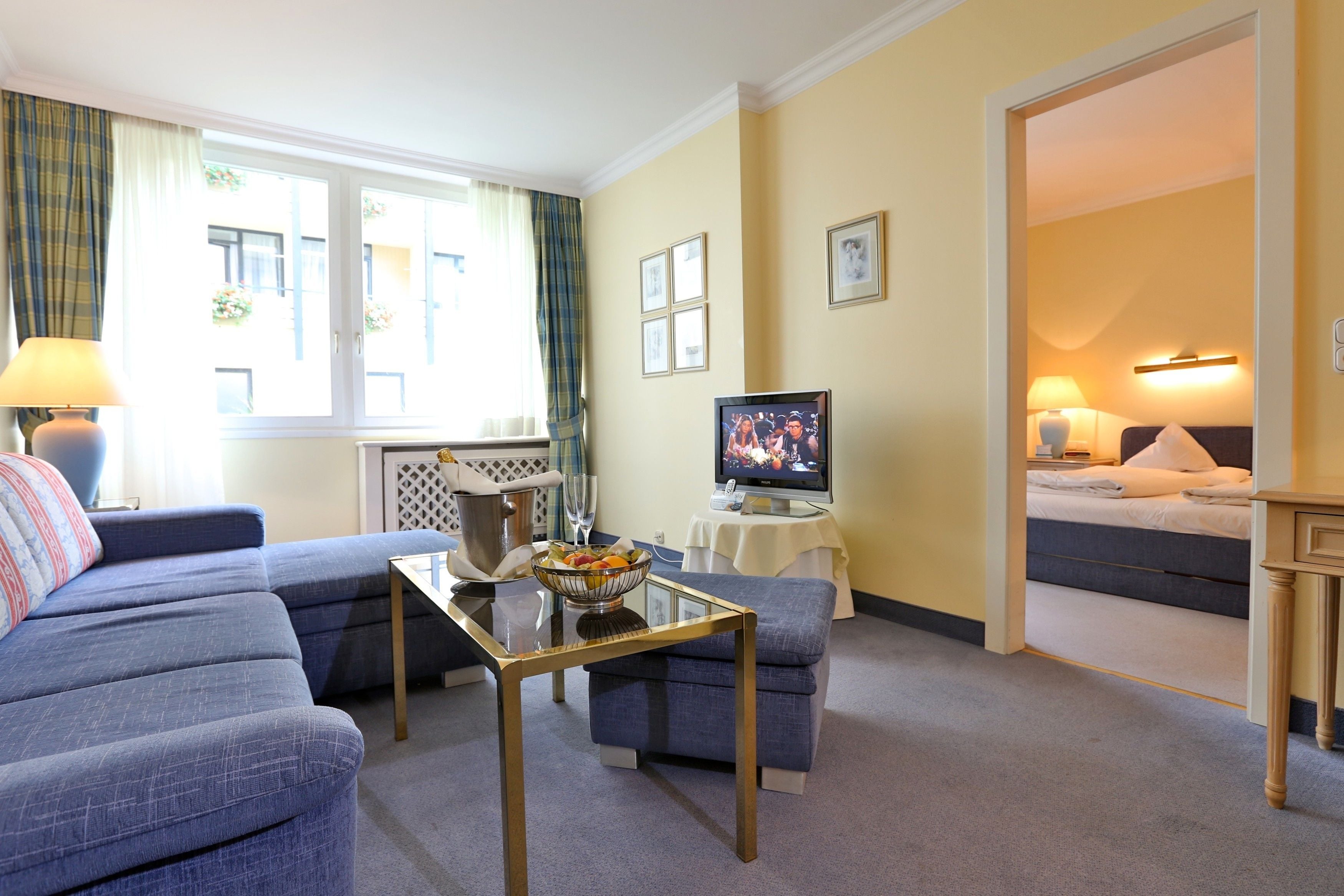 A modern living room with a sofa, a coffee table, and a television. There is a bed in the adjacent room.
