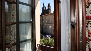 A view through a window with floral curtains, a balcony with a plant, and a building with two towers in the background. - Hostal San Clemente by Pousadas de Compostela (Santiago de Compostela)