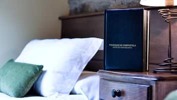 Una cama bien hecha con almohadas blancas y verdes, una mesita de noche de madera con una lámpara, y un libro titulado "POUSADAS DE COMPOSTELA HOTELES CON ENCANTO".