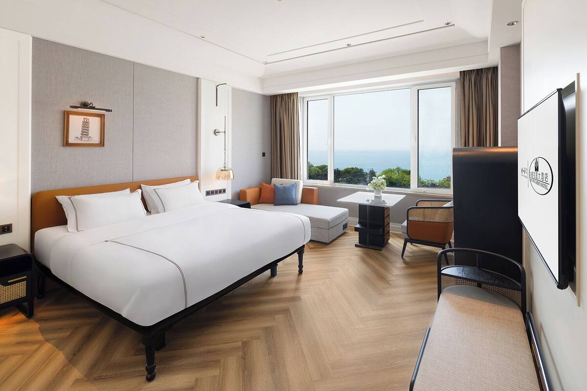 A modern hotel room with a large bed, a seating area with a sofa and armchairs, a small table, and a TV.